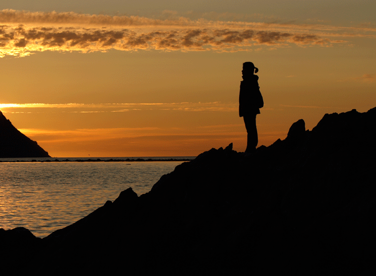 A woman standing on a rocky coastline, silhouetted by a sunset, in Norris Point, Newfoundland, Canada by David P. Lewis